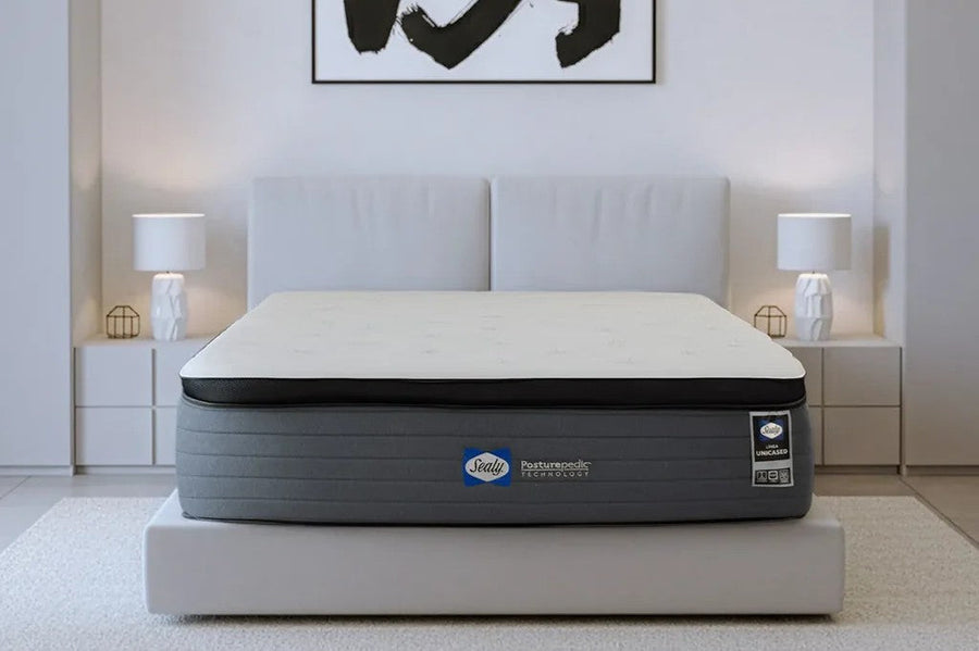 Un colchón Sealy Posturepedic blanco y gris sobre una cama de color blanco en un dormitorio minimalista donde se ven lámparas geométricas y un cuadro colgado.
