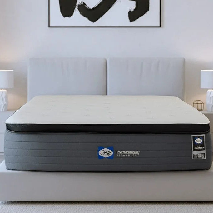 Un colchón Sealy Posturepedic blanco y gris sobre una cama de color blanco en un dormitorio minimalista donde se ven lámparas geométricas y un cuadro colgado.
